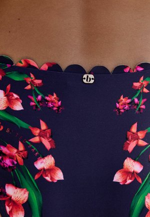 Navy mėlynos audinio kraštas su rausvų ir raudonų gėlių raštu ir mažuoju auksiniu apvaliu prekės ženklo logotipu ant rudos odos fono.