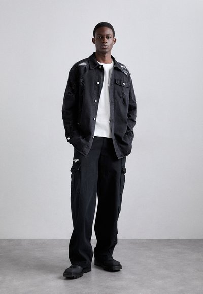 Jachetă oversized din denim negru cu detalii uzate, purtată cu un tricou alb și pantaloni cargo negri largi. Boots negre chunky completează ținuta.
