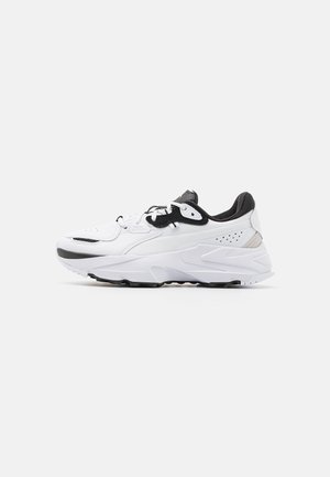 Hvid og sort atletisk sneakers med tyk sål, snøredesign og polstret krave vist fra sideprofil på ensfarvet baggrund.