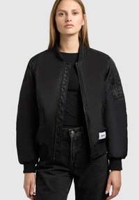 Frau in einer schwarzen gepolsterten Bomberjacke mit Reißverschlusstaschen und gerippten Bündchen über einem schwarzen Shirt und dunklen Jeans, die vor einem grauen Hintergrund steht.