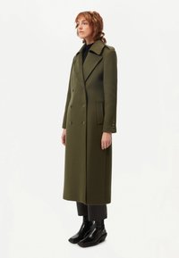 Olivgrön, dubbelknäppt ullrock med stora slag, sidofickor och knappstängning, bärs över svarta byxor och ankelstövlar.