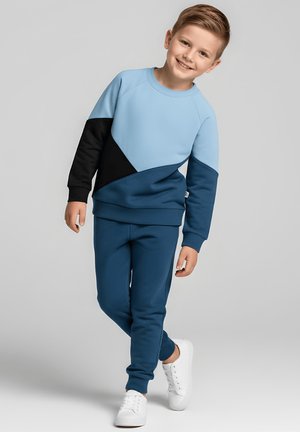 Uśmiechnięty chłopiec w niebiesko-czarnym i jasnoniebieskim kolorowym bluzie i dopasowanych spodniach, idący w białych trampkach na szarym tle.