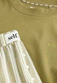 Olive spalvos džemperis su išmegztu apykakle, kuriame yra dryžuotas medvilnės lopas su mygtukais ir mažas išsiuvinėtas logotipas "self."