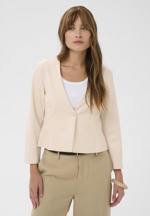 Femeie cu păr brunet lung, purtând un sacou bej peste un top alb și pantaloni bej, stând în fața unui fundal simplu și deschis la culoare.