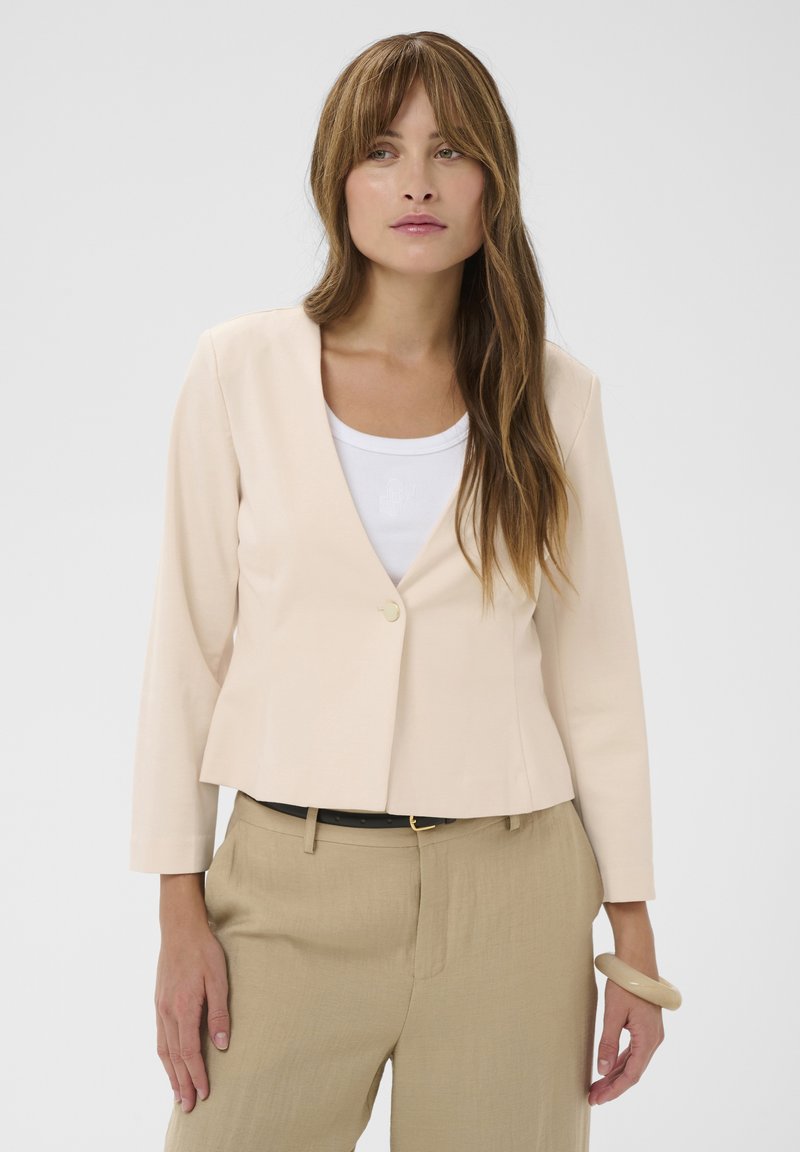Femme aux longs cheveux bruns portant un blazer beige sur un haut blanc et un pantalon beige, debout devant un fond clair uni.