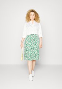 Blusa branca cropped com decote em V, combinada com uma saia floral verde até ao joelho com flores cor-de-rosa. A modelo veste sapatilhas brancas e segura uma bolsa amarela.