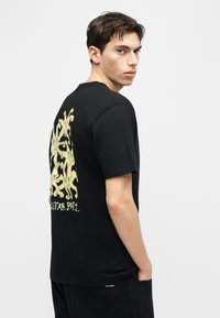 Νέος άνδρας κοιτάζει πάνω από τον ώμο, φοράει μαύρο μπλουζάκι με κίτρινο σχεδιασμό καρτούν αστερία και μερικό κείμενο στην πλάτη, τα χέρια του στις τσέπες μαύρου παντελονιού.