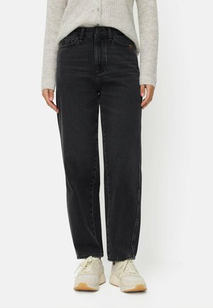Person iført løse sorte denimjeans, hvide sneakers og en lysegrå strikket sweater, stående mod en hvid baggrund.