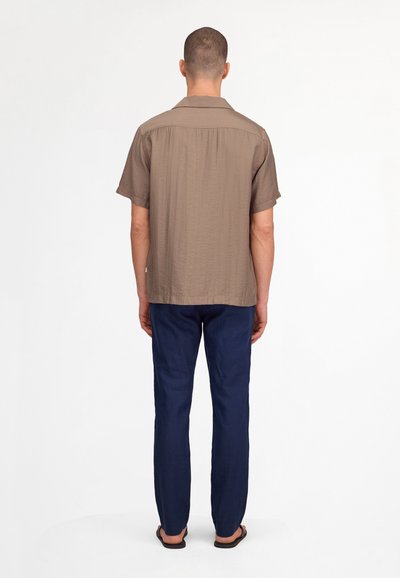 Homme debout, de dos, portant une chemise marron à manches courtes, un pantalon bleu marine et des sandales noires sur un fond clair uni.