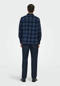 Modra karo s črnimi poudarki, dolgi rokavi in ravno krojem, v kombinaciji s temnimi denim hlačami in črnimi čevlji.