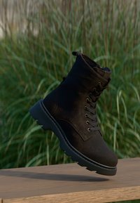 Gheată neagră din piele intoarsă cu finisaj texturat, vârf rotund, design cu șireturi în față și talpă groasă de cauciuc, așezată pe o suprafață din lemn.