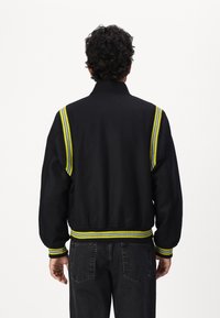 Veste bomber noire avec un col et des poignets côtelés, présentant des rayures jaunes et bleues sur les épaules. Texture douce, coupe décontractée.