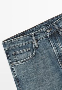 Verbleekte blauwe spijkerbroek tailleband met riemlussen, metalen knoop en voorzakstiksels op een witte achtergrond.