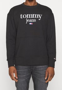 Felpa nera con il testo "tommy jeans" e piccolo logo a bandiera rosso, bianco e blu, indossata con jeans grigio scuro.
