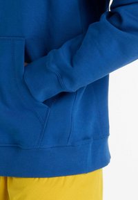 Blå sweatshirt med ribbstickade muddar och fåll, med en framficka. Texturerat tyg; kombinerad med gula shorts för kontrast.