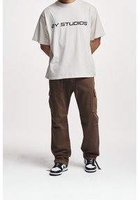 T-shirt di cotone beige chiaro con testo "ZY STUDIOS", abbinata a pantaloni cargo marroni con tasche laterali. Sneakers black and white completano il look.