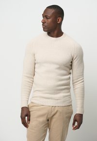 Beige Strickpullover mit Rundhalsausschnitt, strukturiertem Stoff und Rippenbündchen an den Ärmeln sowie am Saum. Kombiniert mit hellen Hosen.