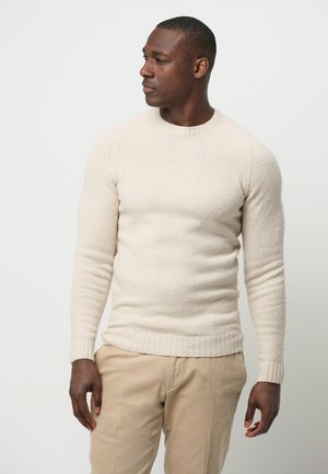 Suéter de punto beige con cuello redondo, tejido texturizado y puños y dobladillo acanalados. Combinado con pantalones de color claro.