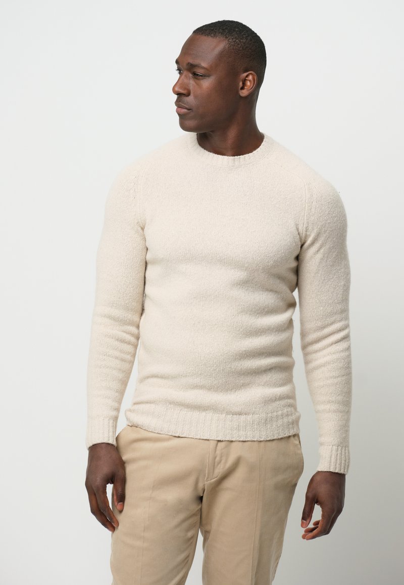 Beige Strickpullover mit Rundhalsausschnitt, strukturiertem Stoff und Rippenbündchen an den Ärmeln sowie am Saum. Kombiniert mit hellen Hosen.