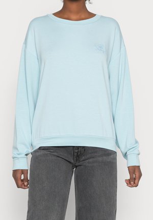 Personne portant un sweat-shirt bleu clair à manches longues avec un logo subtil sur la poitrine et un jean gris foncé, debout devant un fond uni.