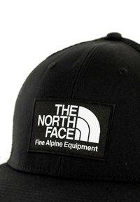 Gorra negra con visera plana, que presenta un parche rectangular blanco con el texto "THE NORTH FACE" y "Equipamiento Alpino Fino."