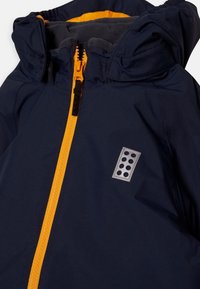 Navy blue jacket with bright orange zipper and hood lining, featuring a reflective rectangular patch with six circular dots on the chest.
