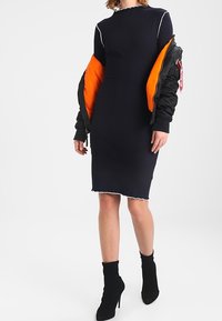 Granatowa sukienka z ribbowanym wzorem, o wysokim dekolcie i falbaniastych brzegach, w zestawieniu z czarną kurtką bomberką z pomarańczowym wnętrzem. Czarne botki za kostkę.