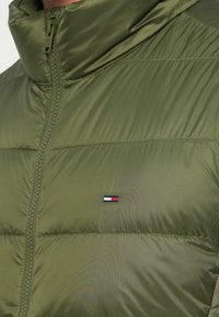 Jachetă pufos verde măsliniu cu guler înalt, având un fermoar și un mic logo brand în roșu, alb și albastru pe partea stângă a pieptului.