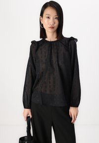 Μαύρη διαφανής μπλούζα με υφή σε ορθογώνια σχέδια, φουσκωτούς ώμους και ελαστικές μανσέτες. Συνδυάζεται με μαύρο παντελόνι και τσάντα.