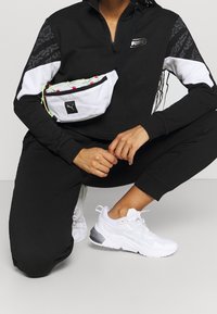 Bolso de pouch blanco con detalles en negro y verde neón, llevado con una chaqueta negra con cremallera y pantalones jogger negros, combinados con zapatillas blancas.
