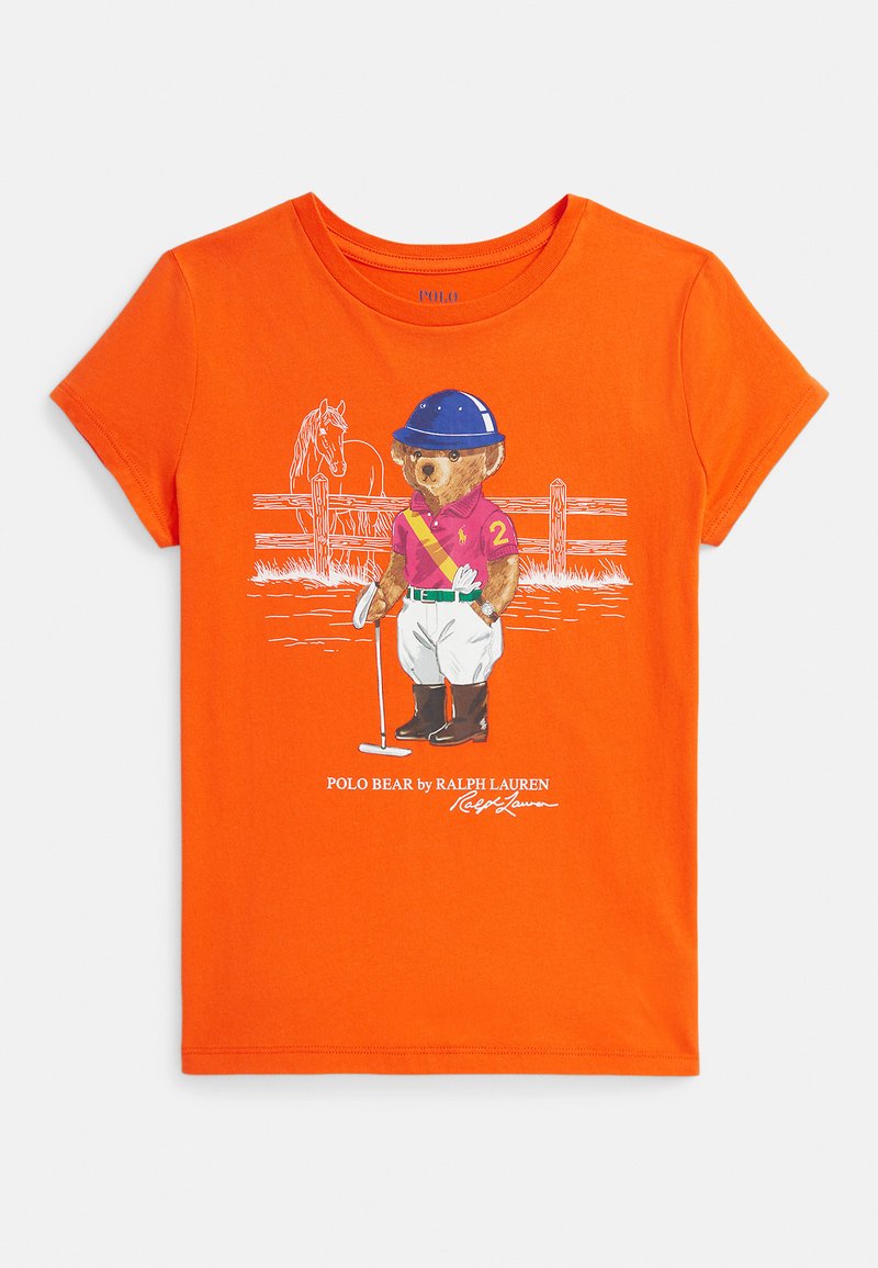 Oranža T veida krekls ar grafiku, kurā attēlots lācis, kas ģērbies polo aprīkojumā un tur rokās āmuru, stāvot priekšā koka žogam un zirgam.