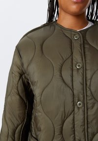 Person wearing an olive green, quilted jacket with wave-like stitching and round buttons over a white shirt, shown from neck to waist.