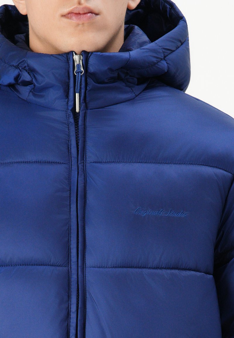 Marineblaue Steppjacke mit einem Reißverschluss vorne, hohem Kragen, gesteppten Paneelen und einem dezenten Logo auf der Brust. Glatte, glänzende Textur.