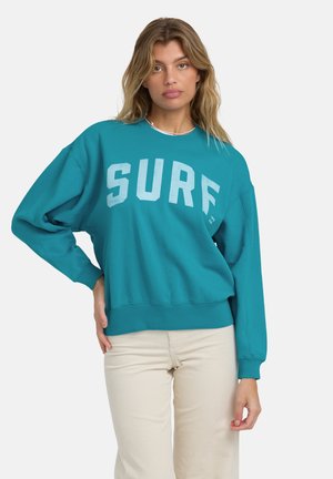 Felpa turchese con stampato audace "SURF" in azzurro chiaro, vestibilità comoda, spalle scese e polsini e orlo a coste. Indossata con pantaloni panna.