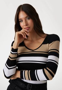 Top long à manches ribbées avec des rayures horizontales noires, blanches et beiges. Design en V avec des manches texturées contrastantes.
