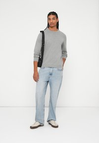 Maglietta grigia a maniche lunghe abbinata a jeans a gamba larga blu chiaro. Borsa nera a tracolla, scarpe da ginnastica bianche con suola marrone. Sfondo semplice.