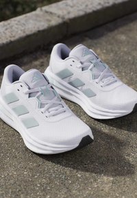 Chaussures de sport blanches avec des rayures et des lacets gris clair sur un pavement extérieur texturé près d’un bord en béton.
