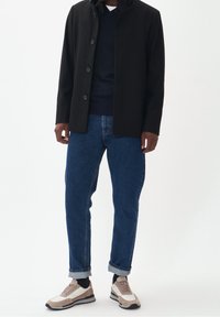 Svart ytterjacka med sex knappar, marinblå tröja under, blå denimjeans och beige/vita sneakers med svarta detaljer.