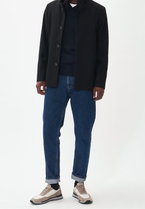 Veste noire à six boutons, pull marine en dessous, jeans en denim bleu et baskets beige/blanc avec accents noirs.