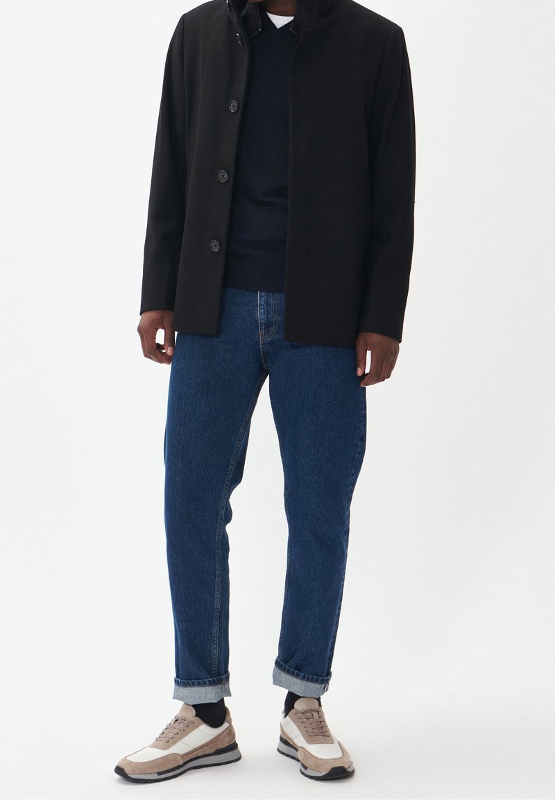 Svart ytterjacka med sex knappar, marinblå tröja under, blå denimjeans och beige/vita sneakers med svarta detaljer.