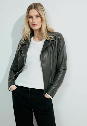 Donkergroene leren motorjack met rits aan de voorkant, gestructureerde stof eronder, en casual zwarte broek, gestyled in een ontspannen pose.