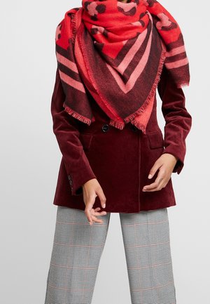Femme portant une grande écharpe à motifs rouges et roses, un blazer en velours côtelé bordeaux et un pantalon à carreaux noir et blanc, debout devant un fond uni.