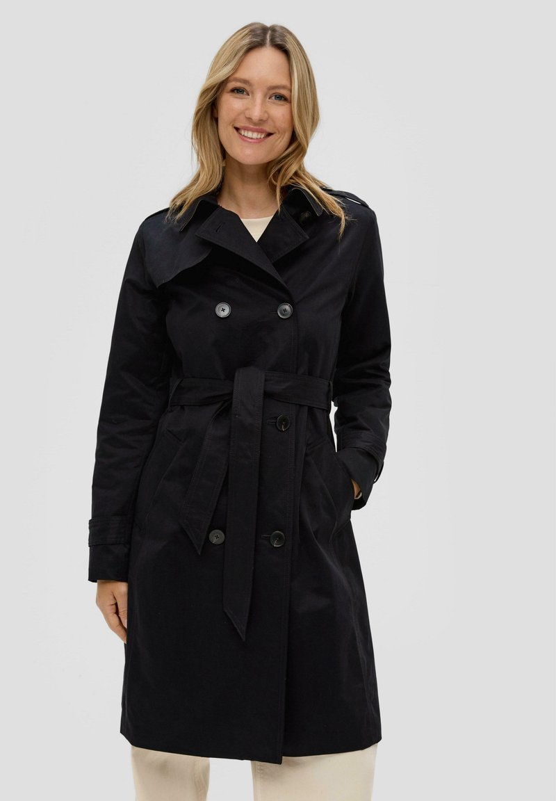 Femme souriante aux cheveux blonds portant un trench-coat noir à double boutonnage avec une ceinture, se tenant devant un fond clair uni.