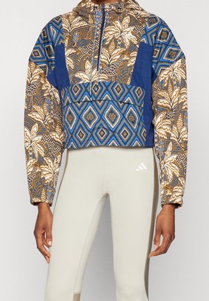 Kort jakke med blå, brune og beige geometriske og blomsteragtige mønstre, båret med lysebeige leggings med hvidt logo.