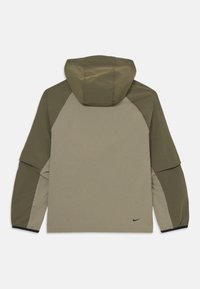 Dvigubos spalvos alyvuogių žalios ir bežinės ilgos rankovės gobtuvas su mažuoju juodu Nike logotipu apatinėje nugaros dalyje, rodomas iš nugaros.