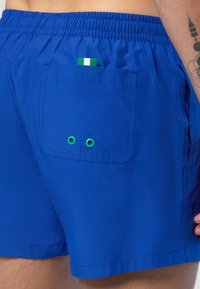 Pantaloni da bagno blu realizzati in tessuto leggero. Presentano una tasca posteriore con occhielli verdi e un piccolo cartellino verde, bianco e rosso. Vita elastica.
