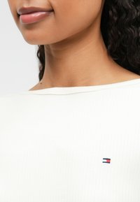 Λευκή ριγέ τοπ με φαρδύ λαιμόκοψη, με μικρό κεντημένο λογότυπο σε κόκκινο, άσπρο και μπλε στο στήθος. Μαλακή υφή.