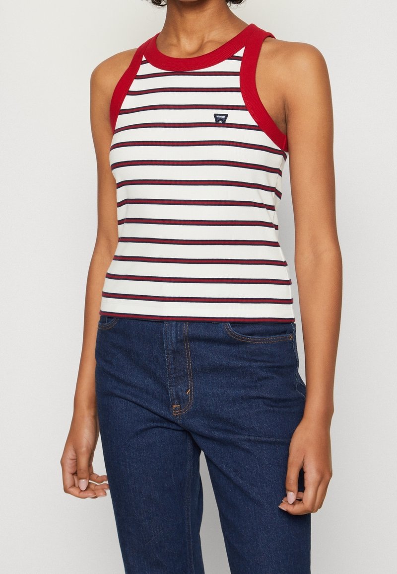 Femme portant un haut blanc sans manches avec une bordure rouge et des rayures horizontales noires, associée à un jean denim bleu foncé.