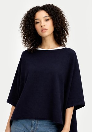 Frau mit lockigem Haar, die ein locker sitzendes marineblaues Oberteil mit weiten Ärmeln und hellblaue Jeans vor einem einfarbigen hellen Hintergrund trägt.