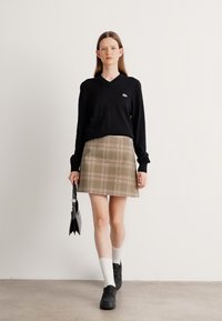 Donna che indossa un top nero a maniche lunghe e una gonna a quadri beige, con in mano una borsa nera, che cammina di fronte a un muro bianco uniforme.
